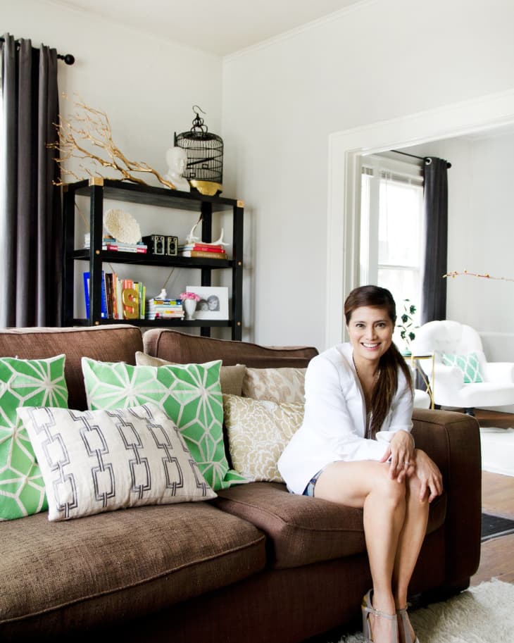 Woman sitting on a brown sofa with green and patterned pillows, next to a black bookshelf with decor items.