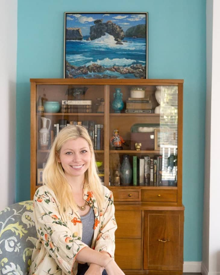 Woman sitting in front of a wooden cabinet with books and decor, turquoise wall, and ocean painting above.