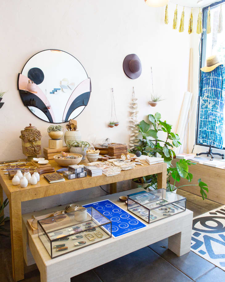 Store interior with wooden tables displaying ceramics, plants, and decor items. A round mirror and hanging hat adorn the wall.