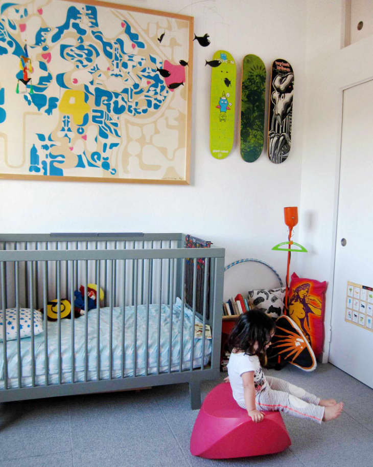 Child sitting on a pink stool in a nursery with a gray crib, colorful wall art, and skateboard decor.