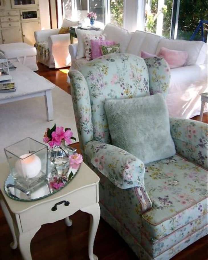 Floral armchair with a gray cushion beside a small table holding a candle and pink flowers in a bright living room.