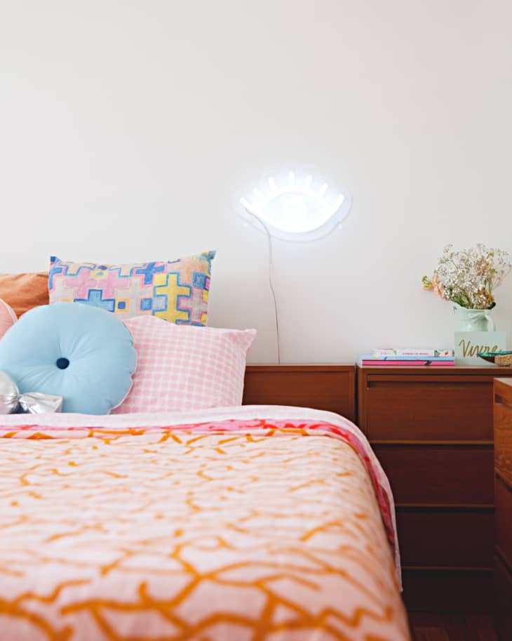 Bedroom with colorful pillows, orange-patterned bedspread, wooden nightstand, and a neon eye wall light.