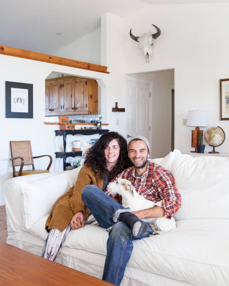 Couple sitting on a white sofa with a dog in a cozy living room, featuring a globe and a bull skull on the wall.