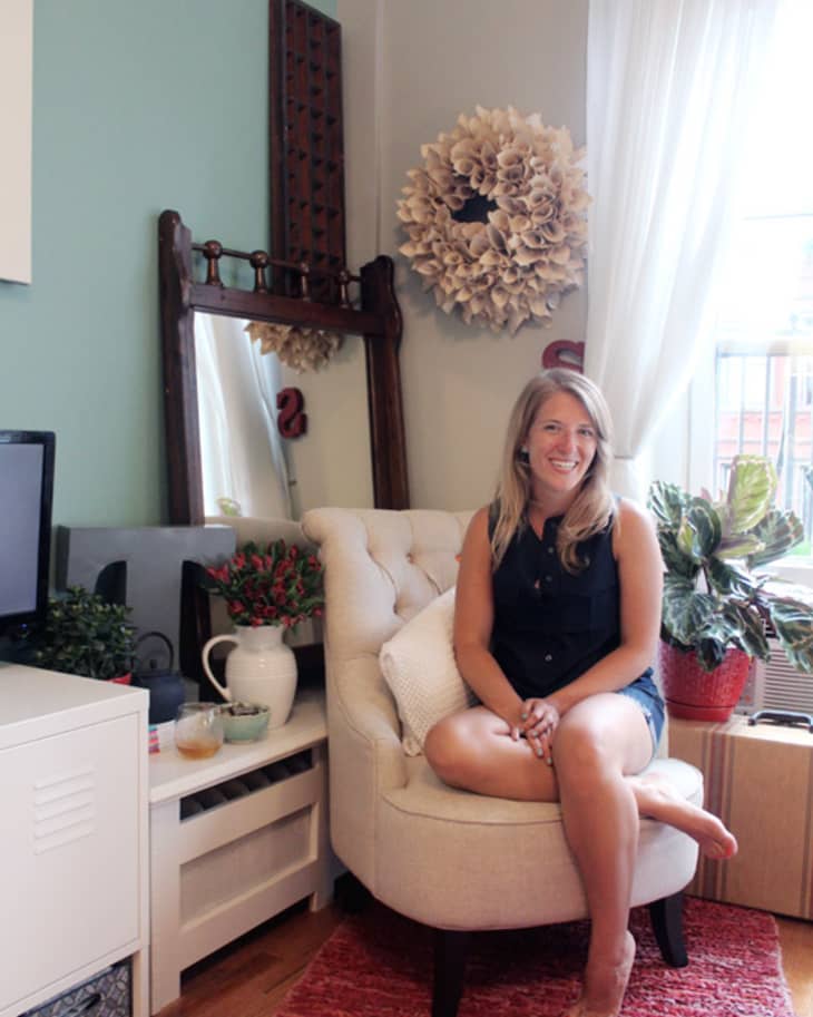 Woman sitting on a cream armchair in a cozy living room with plants, a red rug, and decorative wall mirror.