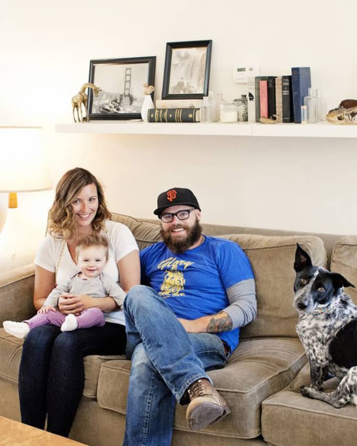 Family sitting on a beige sofa with a baby and a dog, shelf with books and framed photos in the background.