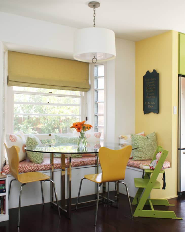 Breakfast nook with yellow chairs, glass table, window seat, colorful cushions, and a vase of orange flowers.