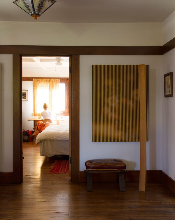 Hallway with wooden floor leading to a bedroom, featuring a bed, a person at a desk, and a large window with curtains.