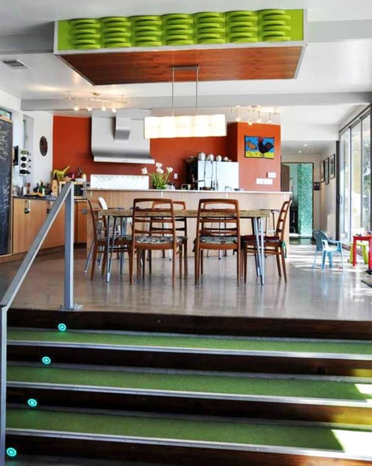 Modern kitchen with orange walls, wooden dining table, chairs, and green ceiling decor, viewed from a staircase.