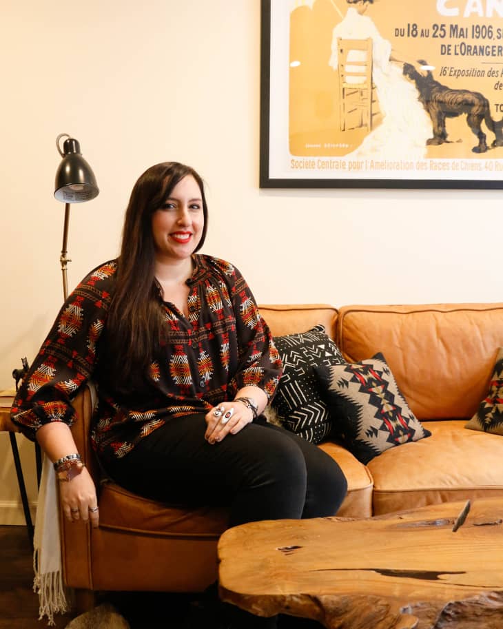 Woman sitting on a tan leather sofa with patterned pillows, next to a wooden coffee table and a floor lamp, under a vintage poster.