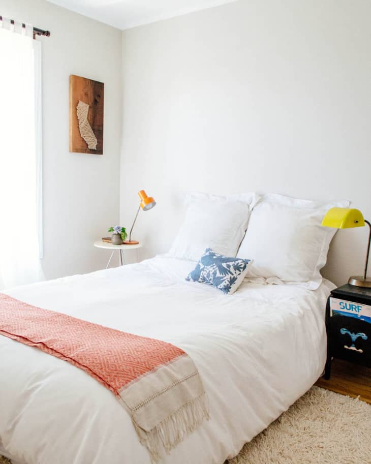 Cozy bedroom with white bedding, orange throw, blue pillow, and surf-themed decor on nightstand.