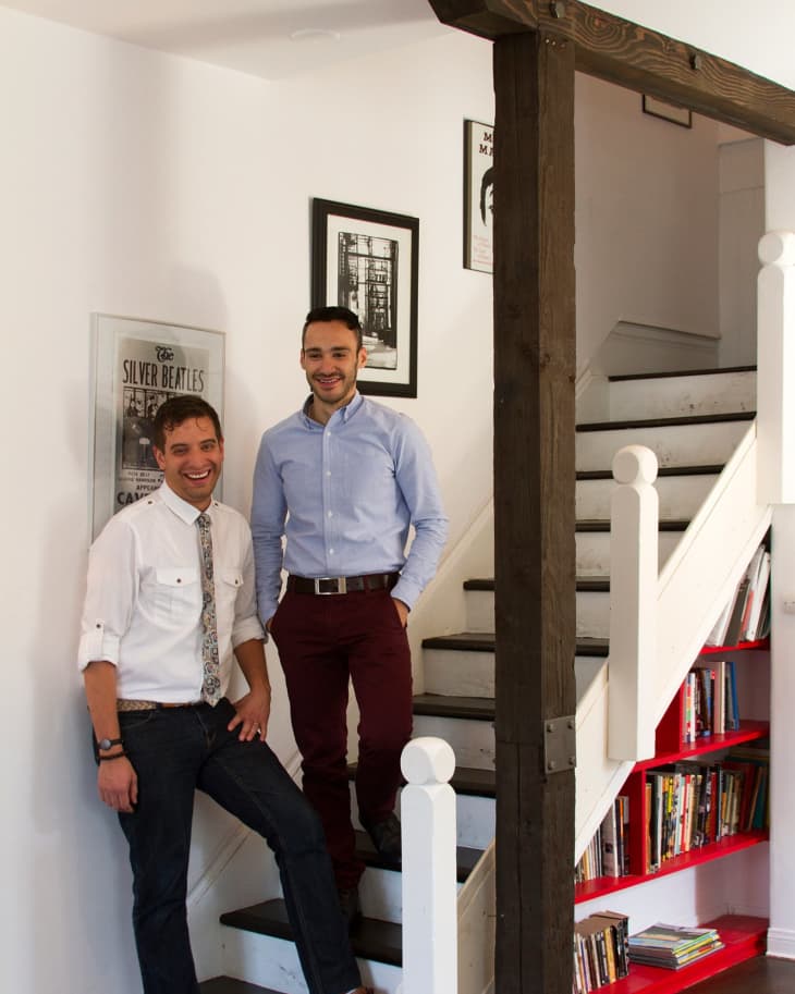 Two men standing on a staircase with red bookshelves, framed posters on the wall.