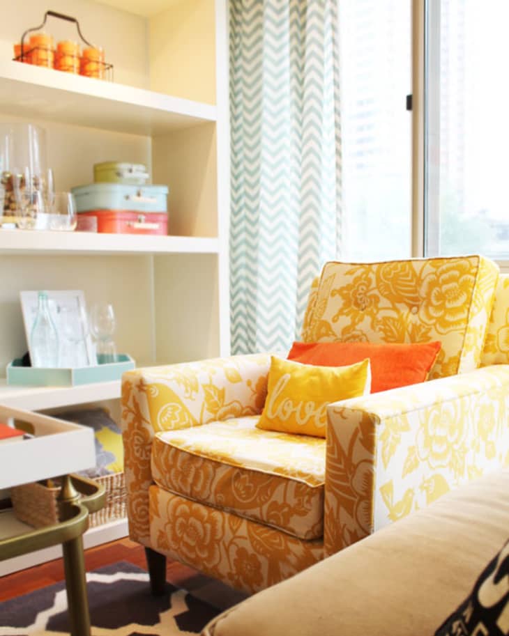 Yellow floral armchair with orange and yellow pillows, near a shelf with colorful boxes and glassware, by a window with chevron curtains.