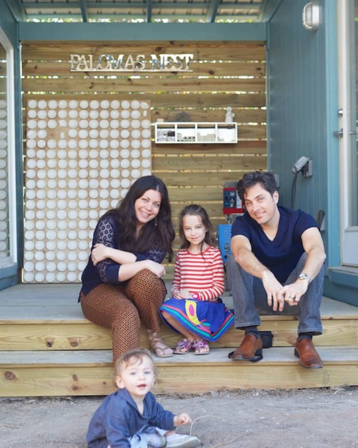Family sitting on wooden steps outside a building with "Paloma's Nest" sign, young child playing in front.
