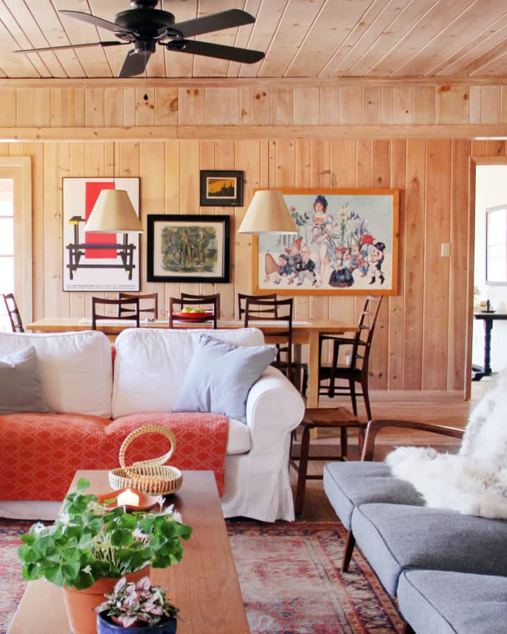 Cozy living room with white sofa, orange throw, wooden coffee table, potted plants, and framed art on wood-paneled walls.