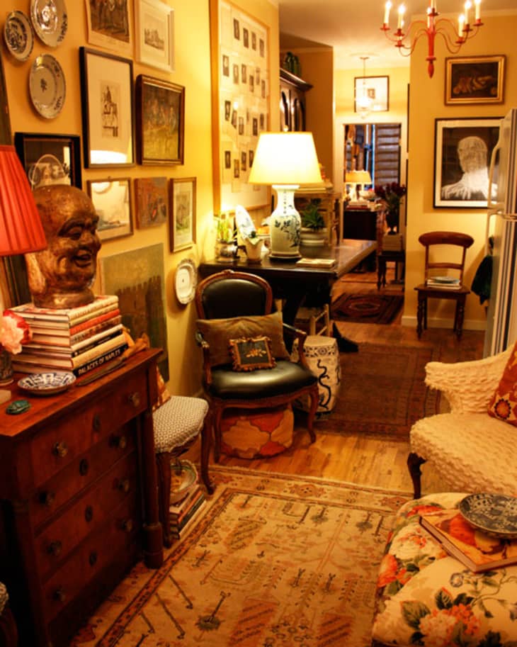 Eclectic hallway with framed art, a wooden bust, floral chair, and a red chandelier.