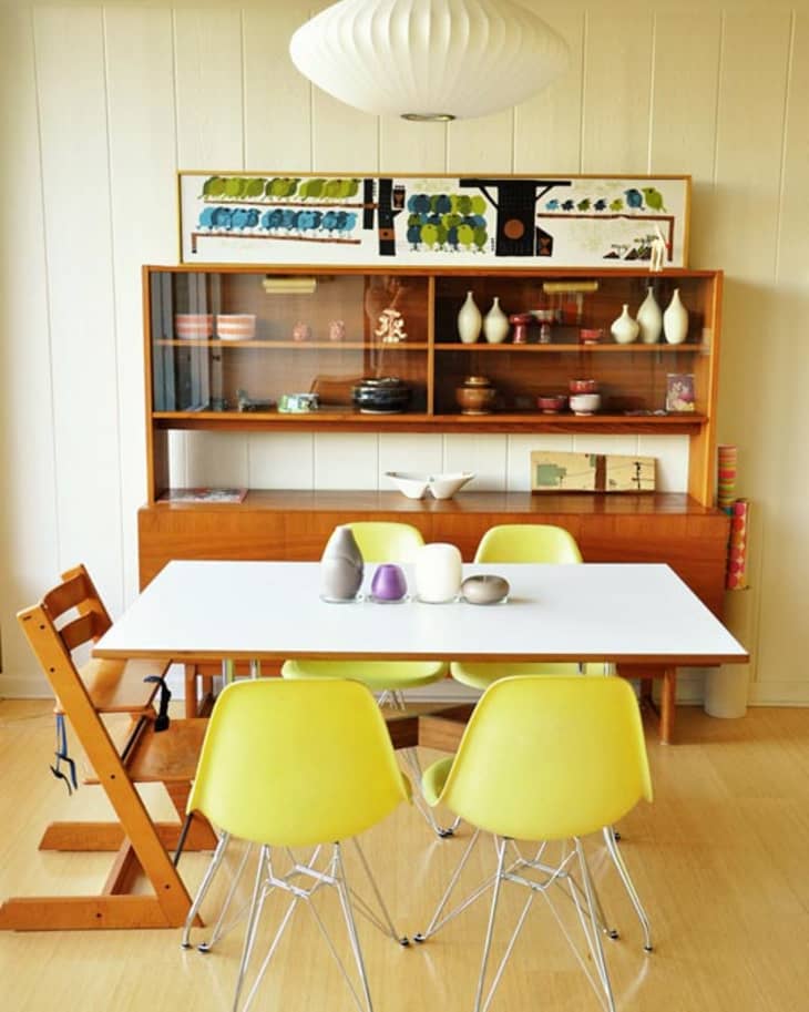 Mid-century dining room with yellow chairs, white table, wooden hutch, and pendant lamp. Decor includes vases and art.