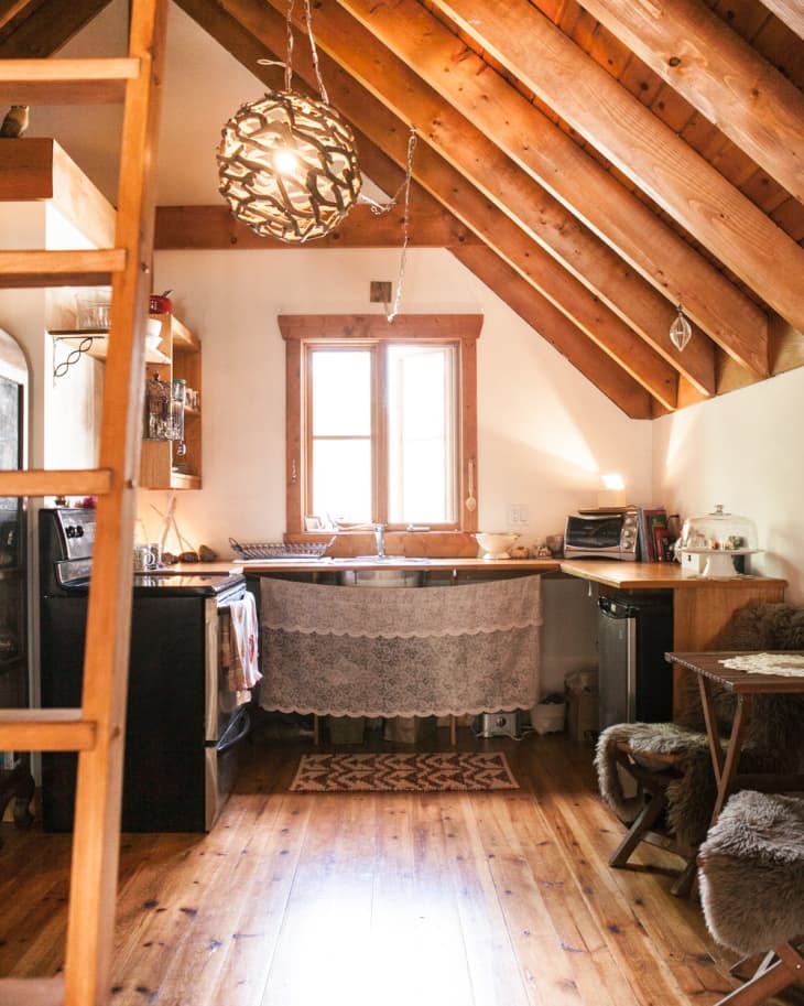 Rustic kitchen with wooden beams, lace curtain under sink, hanging light fixture, and cozy seating area with fur cushions.