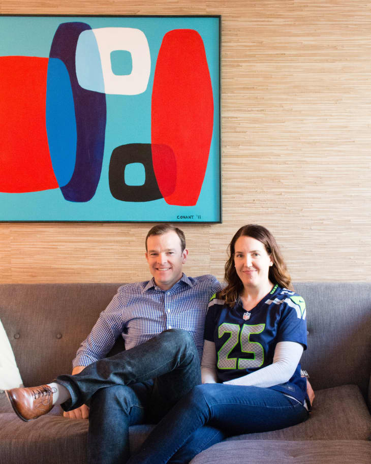 Couple sitting on a gray sofa under a colorful abstract painting, woman wearing a sports jersey.