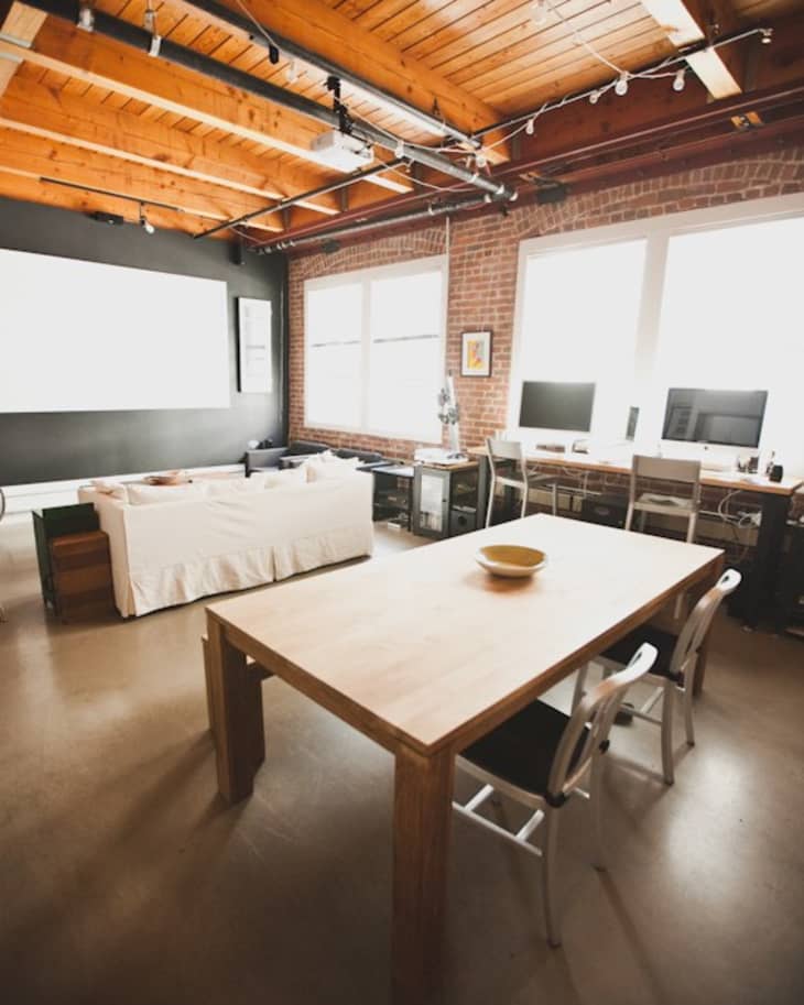 Loft-style office with wooden ceiling, large table, white sofa, and dual computer monitors on desks by windows.