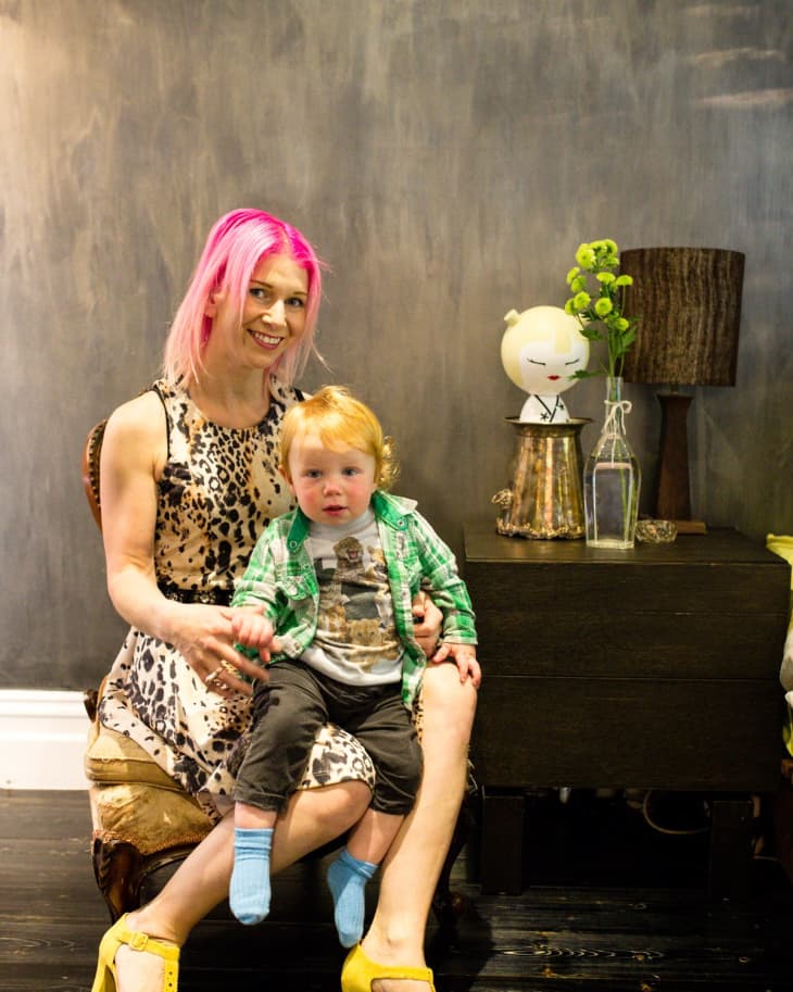 Woman with pink hair and child sitting on a chair, next to a table with a lamp and decorative items.