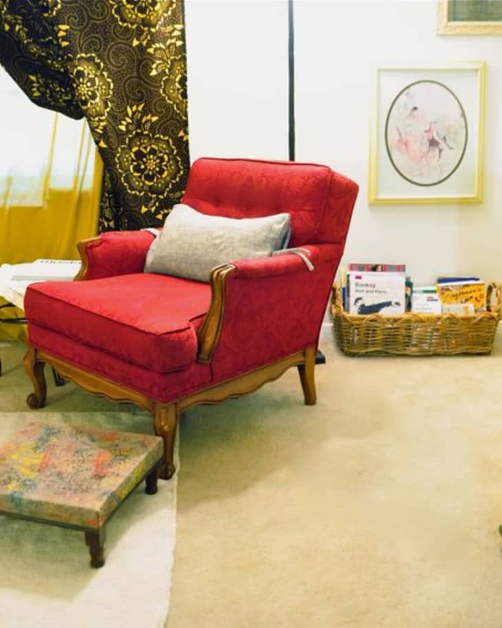 Red armchair with a gray pillow, ornate curtains, a wicker basket with books, and a small footstool in a cozy room.