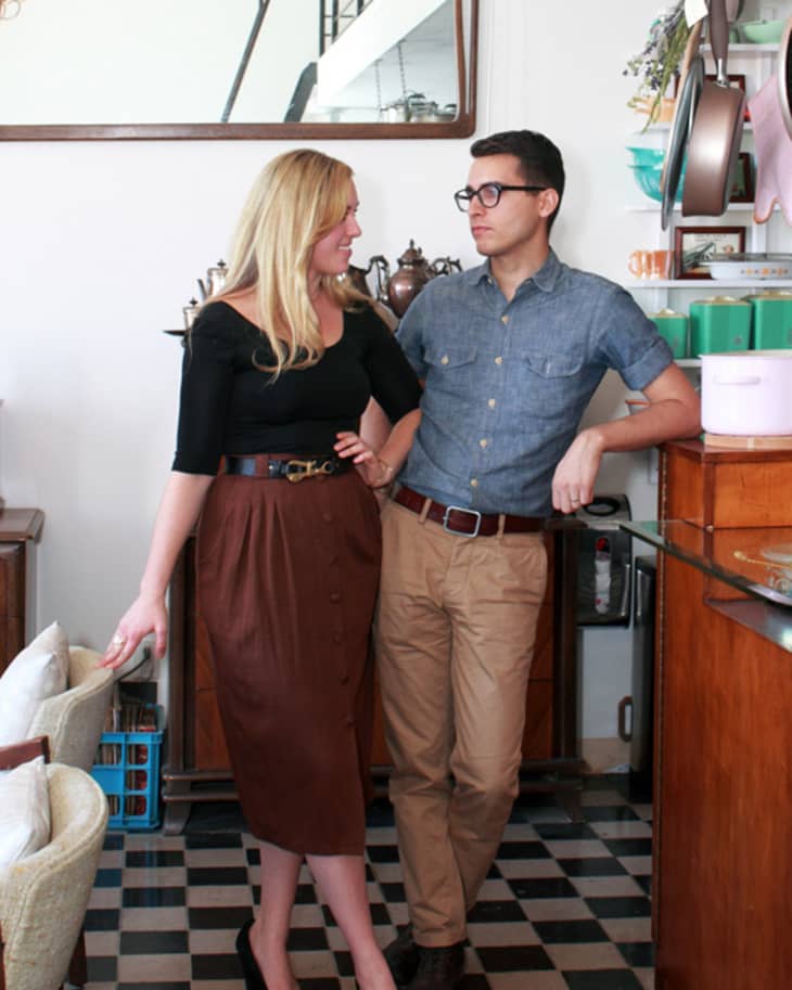 Couple standing in a vintage kitchen with checkered floor, wooden cabinets, and hanging pots.