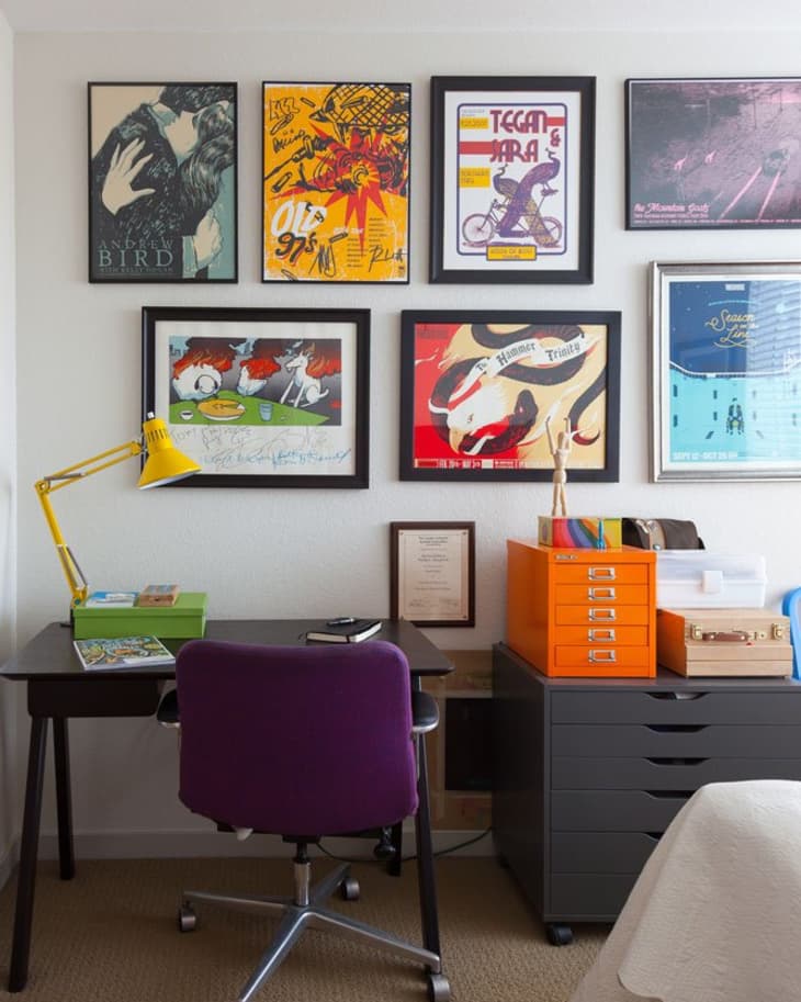 Home office with a purple chair, black desk, yellow lamp, and colorful framed posters on the wall.