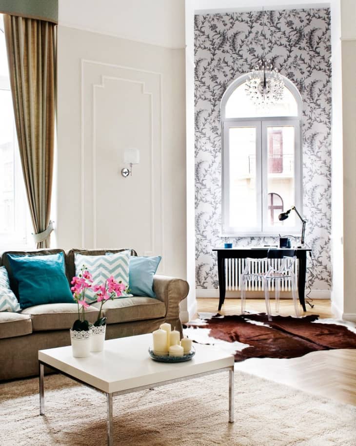 Living room with beige sofa, teal pillows, white coffee table, pink orchids, and a desk by a patterned wallpapered wall.