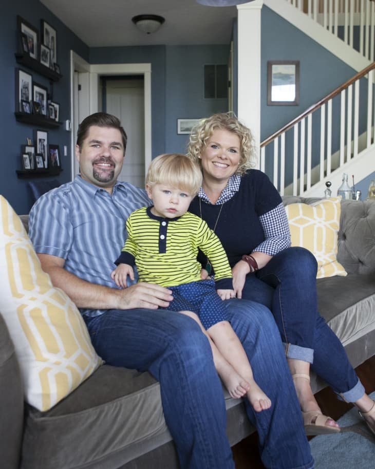 Family sitting on a gray sofa in a living room with blue walls, yellow patterned cushions, and staircase in the background.