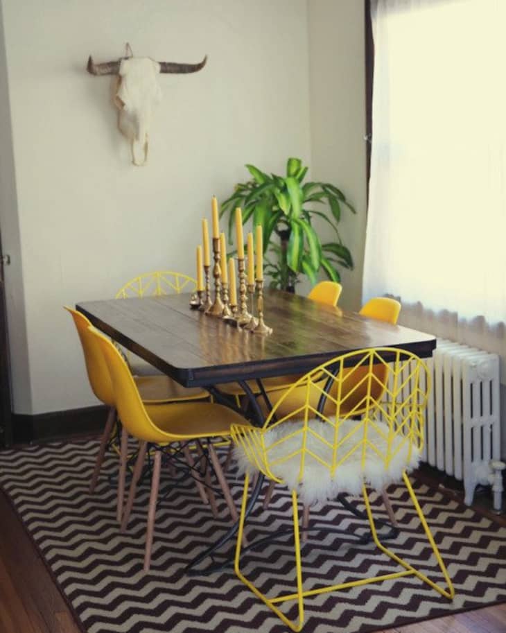 Dining room with yellow chairs, wooden table, gold candlesticks, plant, and a wall-mounted skull above a zigzag rug.