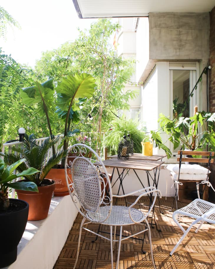 Small balcony with potted plants, white metal chairs, and a wooden table under a covered ceiling.