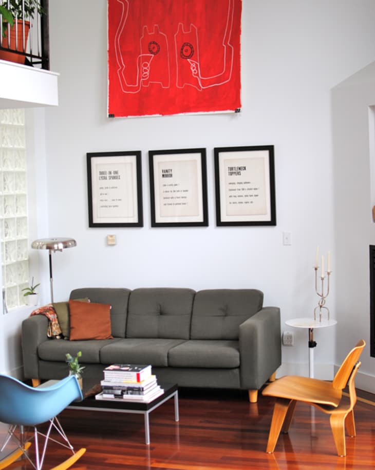 Living room with gray sofa, colorful art, blue rocking chair, wooden chair, and framed prints on white walls.