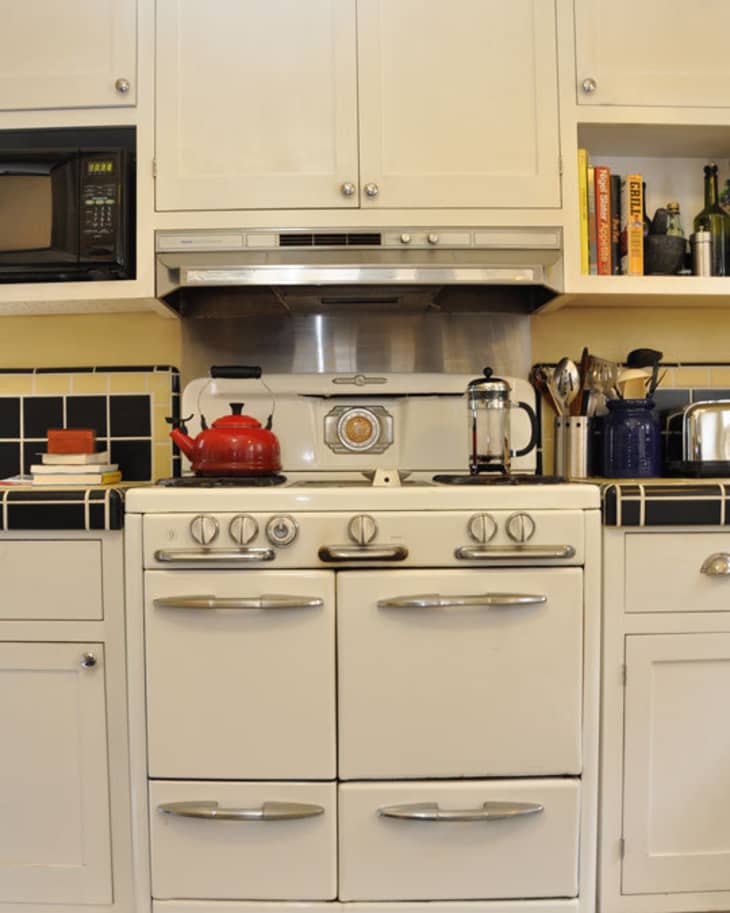 Vintage white stove with red kettle, French press, and utensils on a black-tiled kitchen counter.
