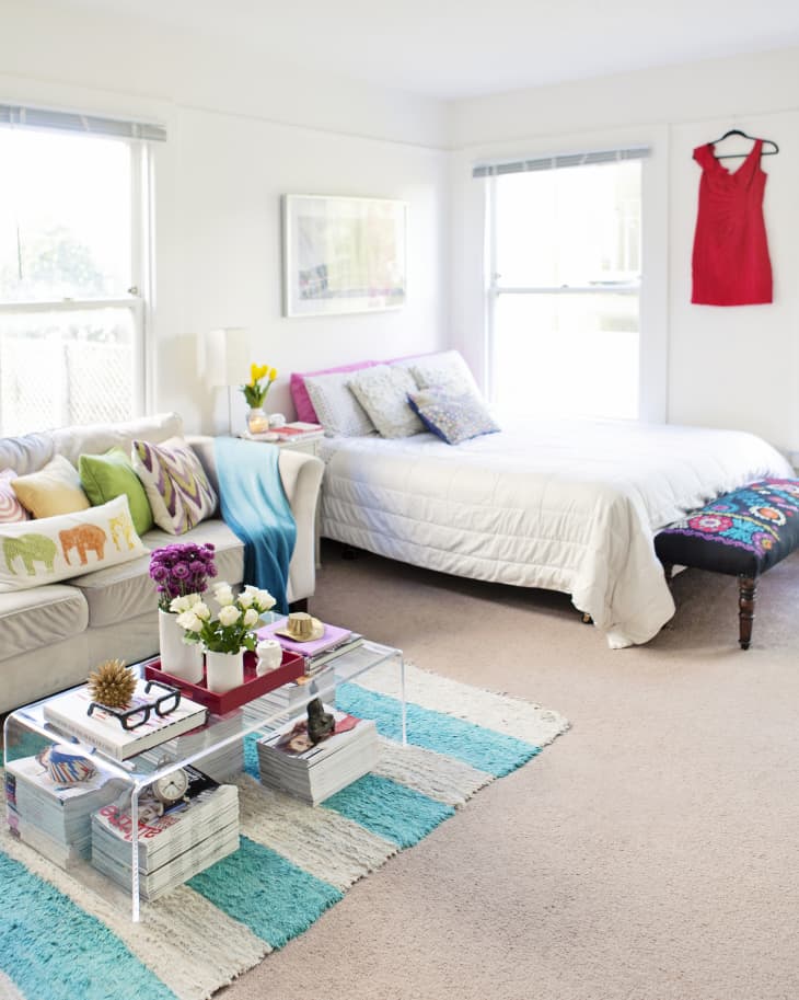 Bright bedroom with a white bed, colorful pillows, glass coffee table, and a red dress hanging on the wall.