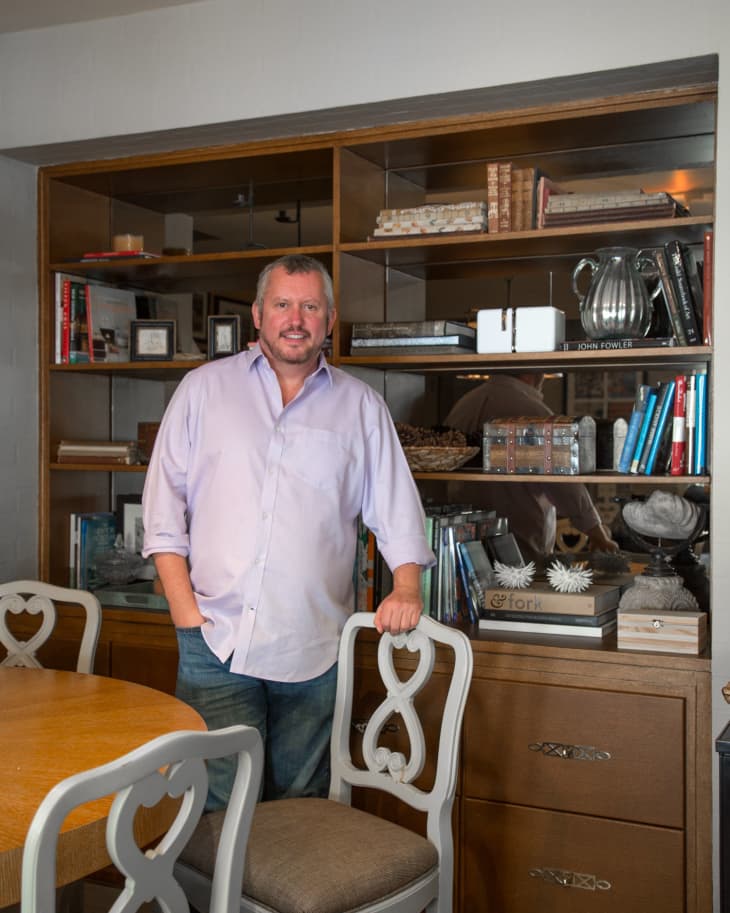 Man in a light purple shirt standing by a wooden bookshelf filled with books and decor, next to a dining table with ornate chairs.