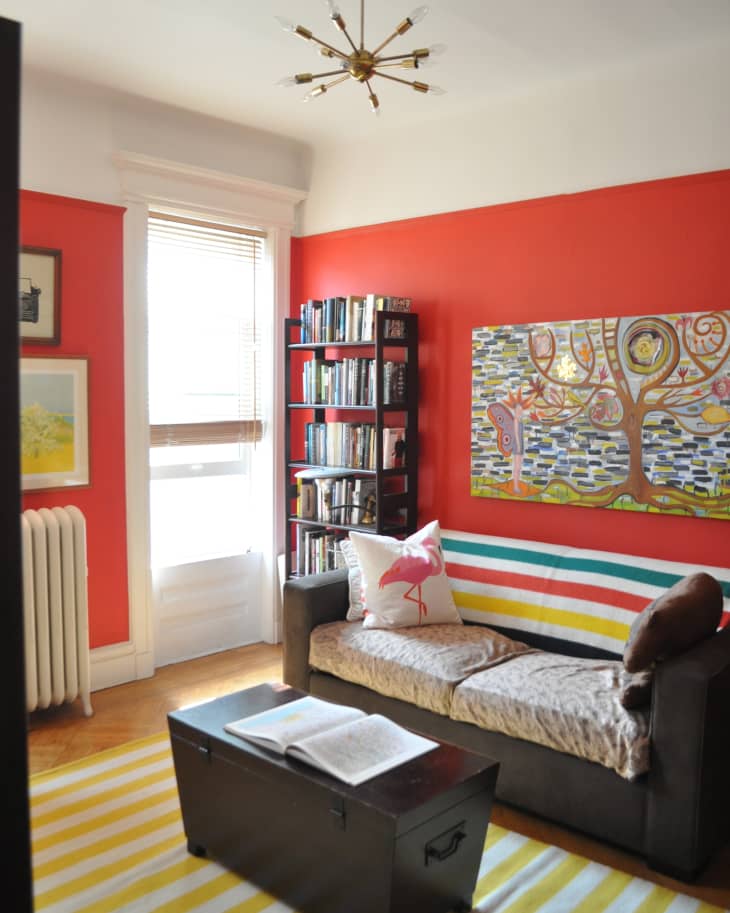 Cozy living room with red walls, striped rug, dark sofa, flamingo pillow, bookshelf, and colorful tree artwork.