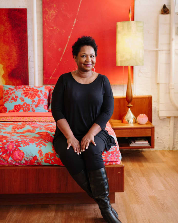 Woman in black outfit sitting on a bed with floral bedding, red abstract art, and a vintage lamp in the background.