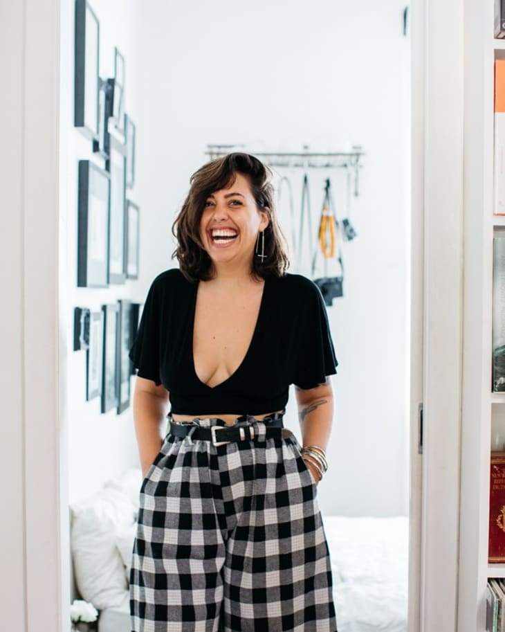 Woman in black top and checkered pants smiling in a room with framed photos and wall hooks.