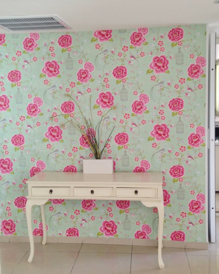 Floral wallpaper with pink roses behind a white console table and vase with branches.