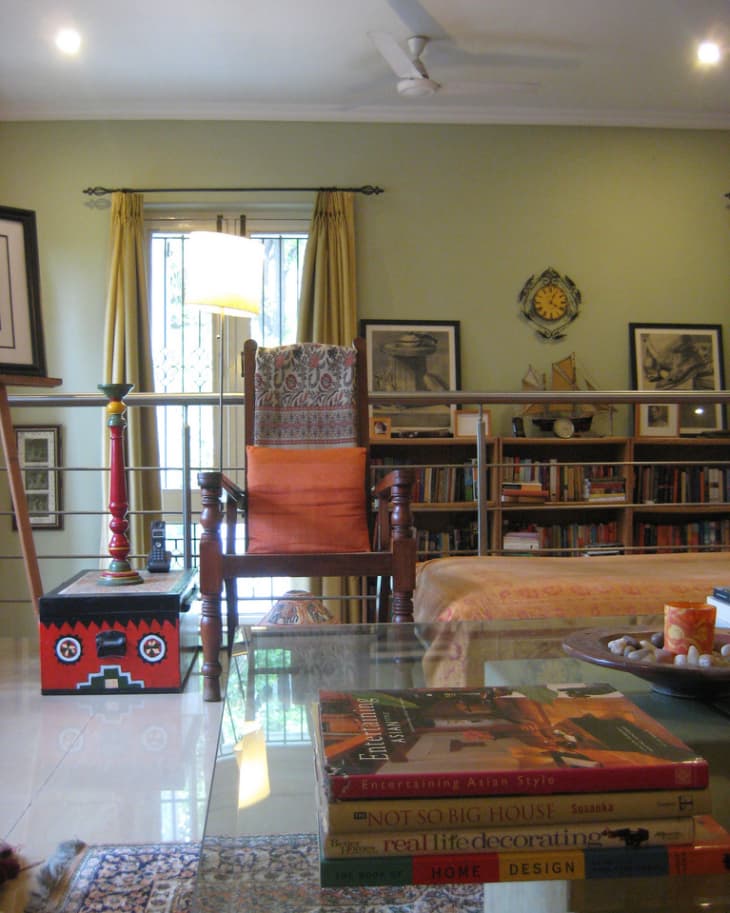 Living room with a wooden chair, glass coffee table, books, and a colorful box, surrounded by framed art and a bookshelf.