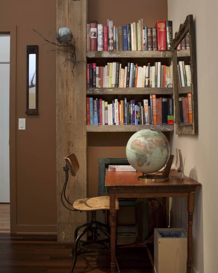 Cozy study nook with wooden desk, vintage chair, globe, and bookshelves filled with colorful books against a brown wall.