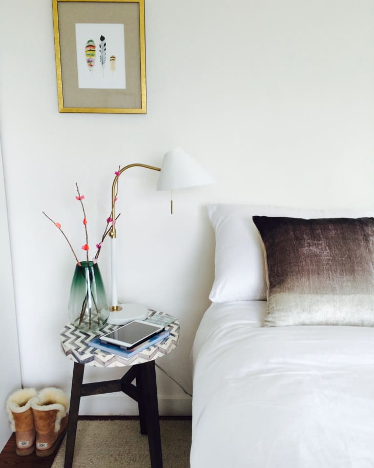 Bedroom with a white bed, gradient pillow, side table with a lamp, vase, and tablet, framed feather art above.