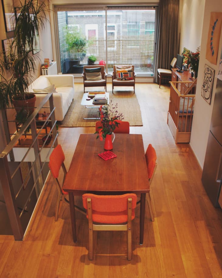 Open-plan living and dining area with wooden table, orange chairs, white sofa, and large window overlooking a balcony.