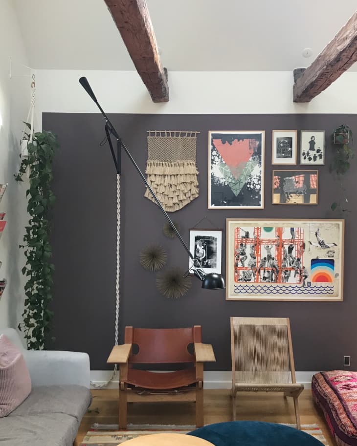 Living room with eclectic wall art, leather chair, woven seat, and hanging plants under exposed wooden beams.