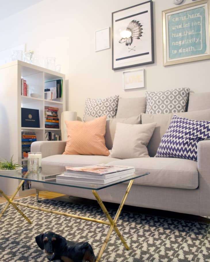 Living room with a beige sofa, patterned cushions, glass coffee table, bookshelf, and wall art.