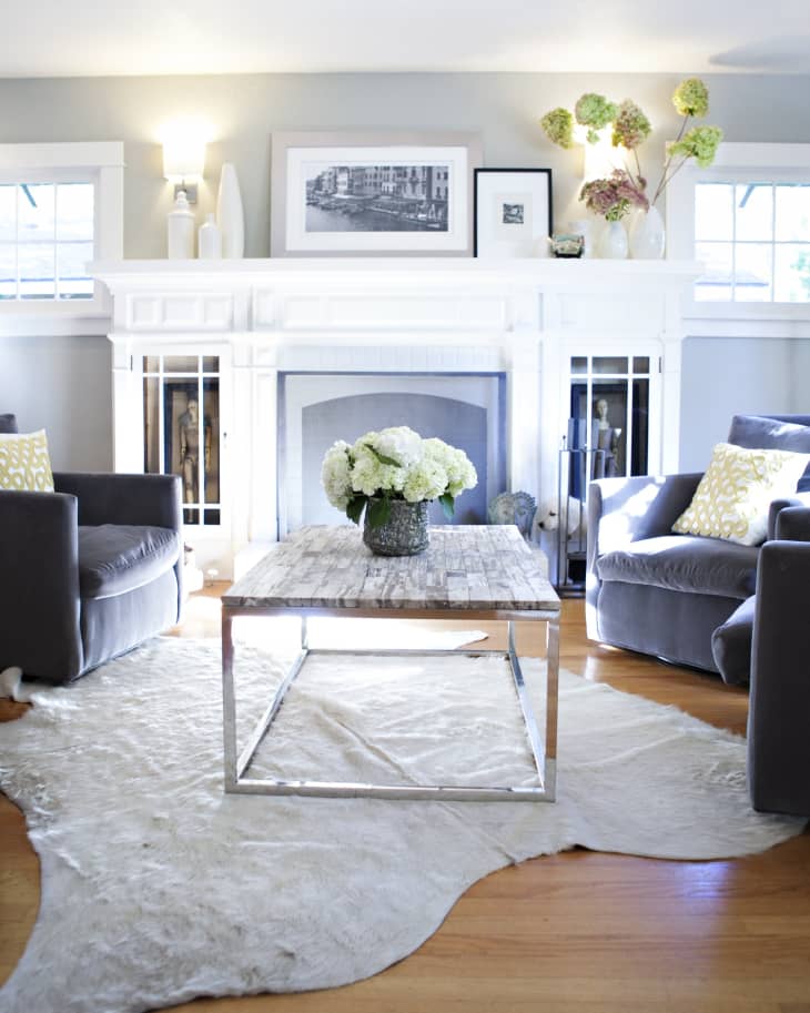 Living room with gray armchairs, white fireplace, rustic coffee table, and a vase of hydrangeas on a cowhide rug.