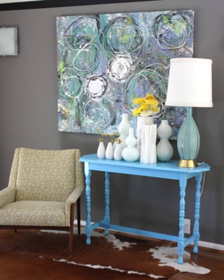 Gray wall with abstract art, blue console table, white vases, yellow flowers, and a beige armchair on a cowhide rug.