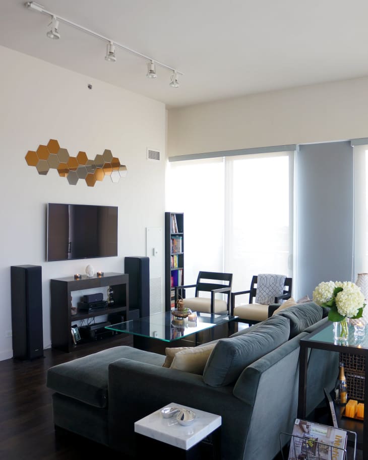 Modern living room with dark sofa, glass coffee table, wall-mounted TV, hexagonal mirrors, and large windows.