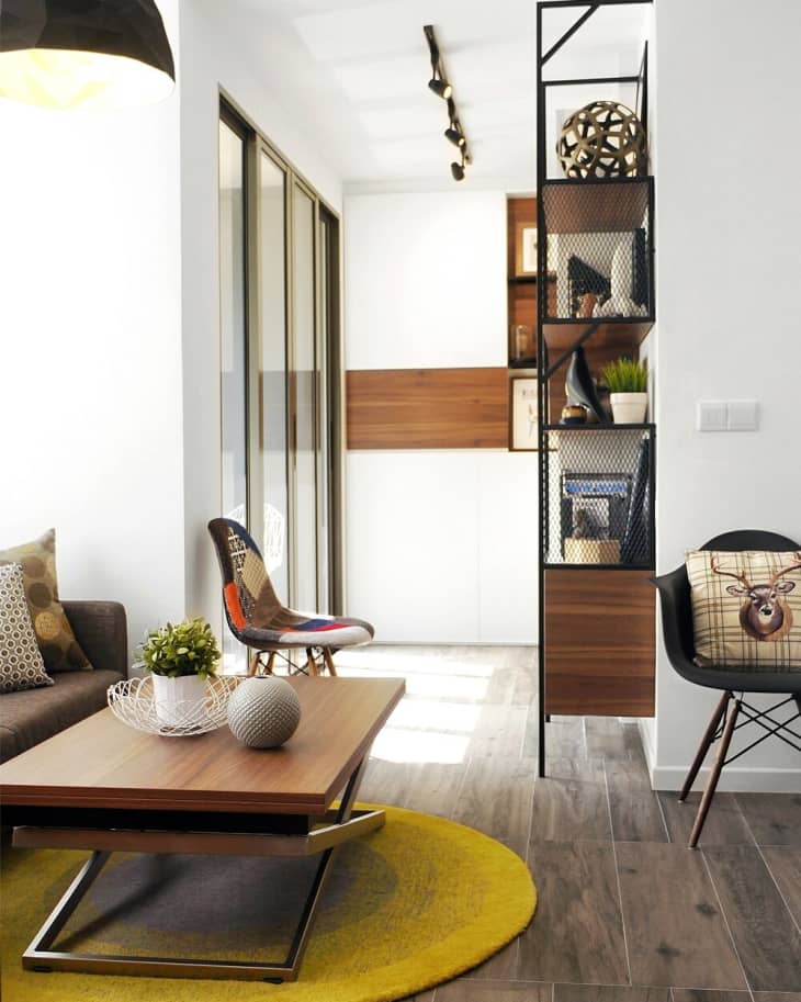 Modern living room with a wooden coffee table, yellow rug, eclectic chairs, and decorative shelving.