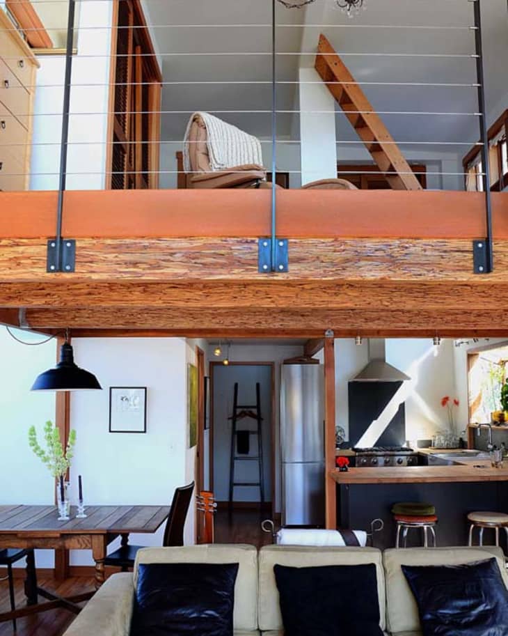Loft-style living room with beige sofa, black cushions, open kitchen, dining table, and upper mezzanine with railing.